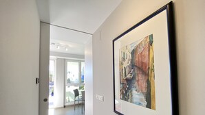 A hallway with a framed painting of a street scene, a door, and a glimpse of a dining area with a table and chairs.