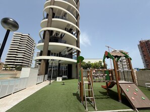 A playground with climbing structures and a slide, surrounded by a building with multiple balconies and a clear sky. - Alq 200 Infinium II 1 (Calpe)