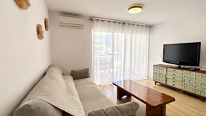 Ein Wohnzimmer mit einer Couch, einem Couchtisch, einem Fernseher auf einem Schrank und einem großen Fenster mit Vorhängen.