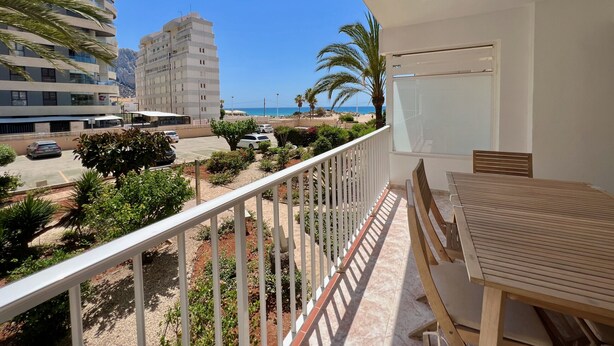 Ein Balkon mit Tisch und Stühlen, der auf einen Parkplatz mit Autos und Grünflächen blickt, sowie eine Aussicht auf einen Strand und Gebäude in der Ferne.