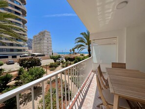 A balcony with a table and chairs, overlooking a parking area with cars and greenery, and a view of a beach and buildings in the distance. - Alq 217 Peñon de Ifach 1 1 (Calpe)