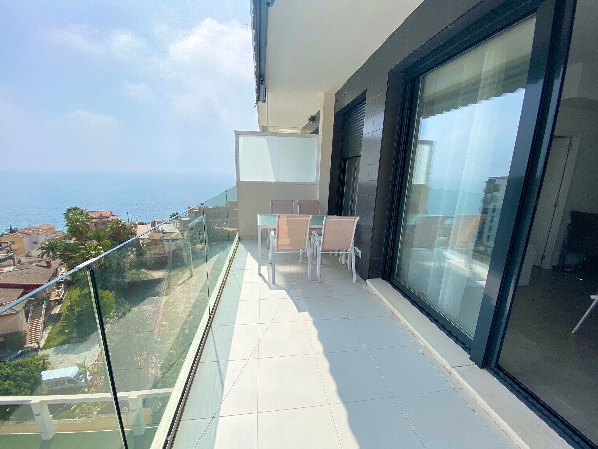 Een balkon met een glazen hekwerk, een tafel met stoelen, en uitzicht op zee en gebouwen.
