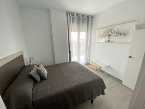 A bedroom with a bed, a small desk, a chair, a window with curtains, and a wall-mounted shelf with a plant. - Alq 229 Calpe Beach (Calpe / Calp)