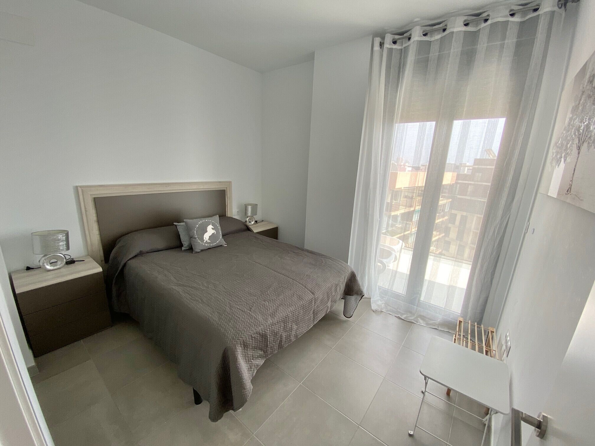 A bedroom with a bed, nightstands, a lamp, a window with curtains, and a view of buildings outside.