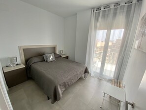 Une chambre à coucher minimaliste, avec un lit, des tables de chevet, une lampe et une vue sur la ville à travers des rideaux transparents.