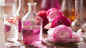 Eine Glasflasche mit rosafarbenem Inhalt, ein Glasgefäß mit Rosenblüten und eine Schale mit rosa Blüten auf einer Holztischplatte.