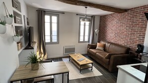 Ein modernes Wohnzimmer mit einer braunen Ledersofa, einem Holztisch und einem Essbereich mit kleinem Tisch und Stühlen. Zwei Fenster mit Vorhängen, eine Ziegelwand und eine Decke mit sichtbaren Balken.