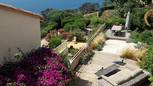 Una terraza con mobiliario exterior, rodeada de abundante vegetación y vistas al mar.