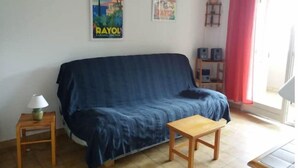 Un salón con un sofá azul, una mesita de madera y una pequeña mesa. Hay carteles en la pared y una cortina roja en la ventana.