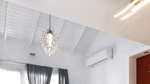 Una sala de estar moderna con sofá, mesa de comedor y sillas. Se observa una araña, una pequeña mesa con una planta y una ventana con cortinas.