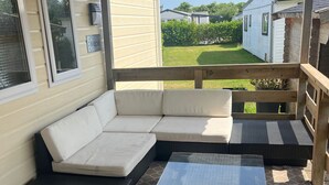 A covered outdoor seating area with a sectional sofa and a coffee table, wooden deck flooring, and a view of a residential neighborhood. - CS 53 - Vakantiepark Callassande (Callantsoog)
