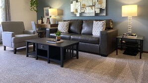 Un salon moderne avec un canapé en cuir foncé, deux fauteuils, une table basse et une table d’appoint avec une lampe. Un élément décoratif est accroché au mur, au-dessus du canapé.