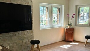 Amplia habitación con pared de piedra, suelo de madera, televisor montado y dos ventanas con vistas a una zona montañosa.