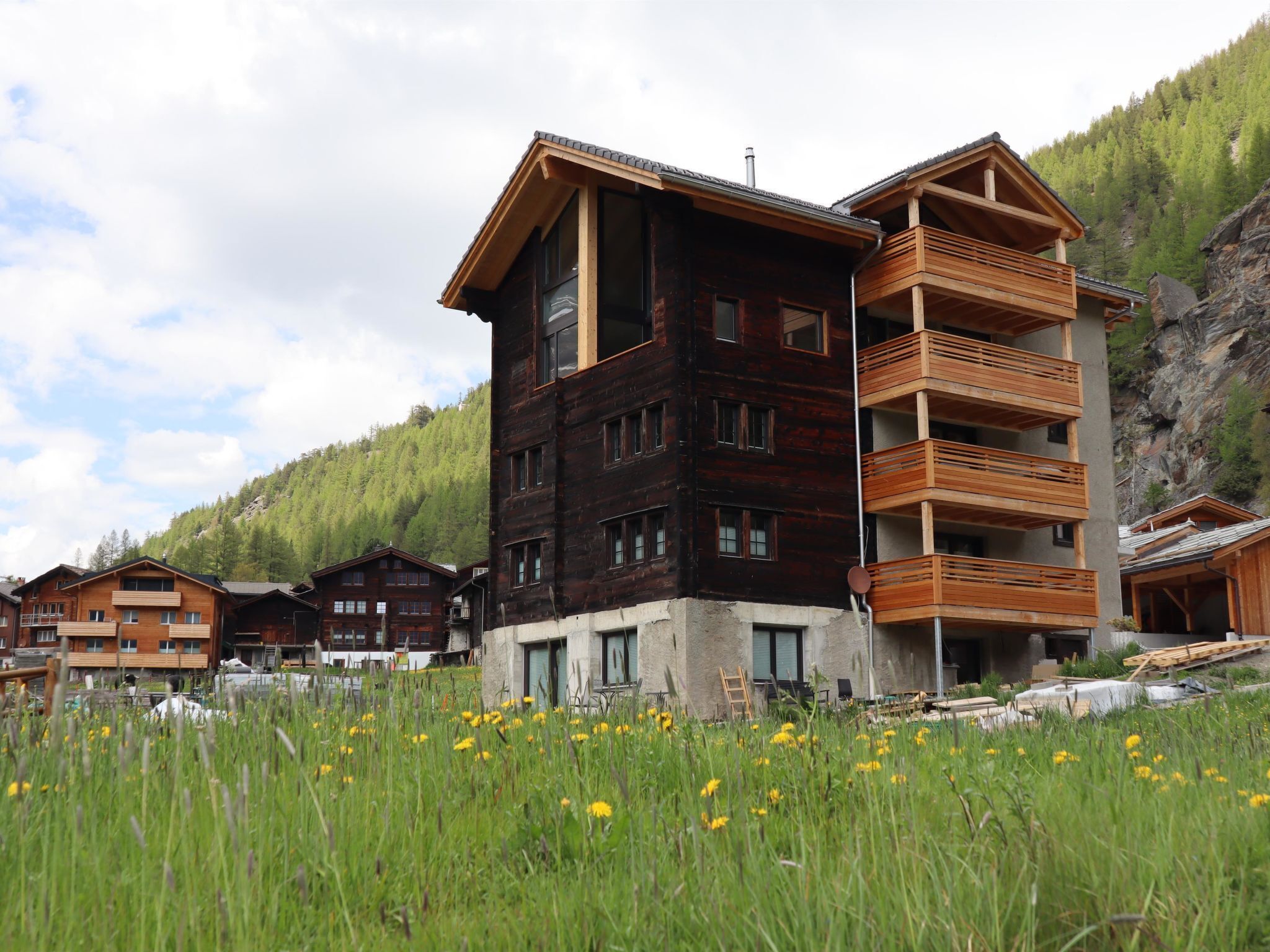 Un bâtiment en bois à plusieurs étages, doté de balcons, entouré d’un champ verdoyant et adossé à un paysage montagneux.