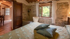 A bedroom with a stone wall, a wooden ceiling, a bed with floral bedding, a window with curtains, and a chair.