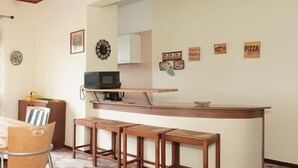 A compact kitchen with a microwave, a coffee maker, and a small refrigerator. There are three wooden stools and a table with a chair. The floor is tiled with a mosaic pattern.