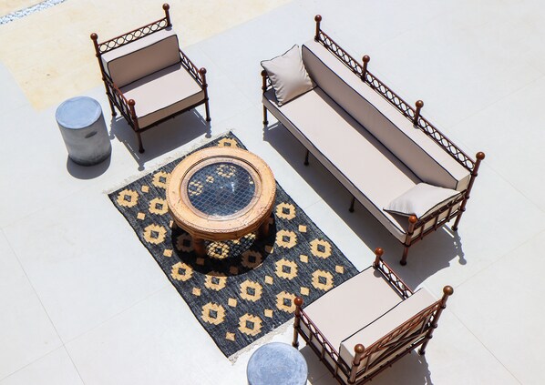 A sunlit outdoor seating area with a round table, two chairs, a sofa, and a small stool.