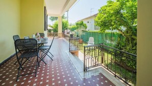 Een balkon met betegelde vloer, een tafel met stoelen en uitzicht op een groene hek en bomen.