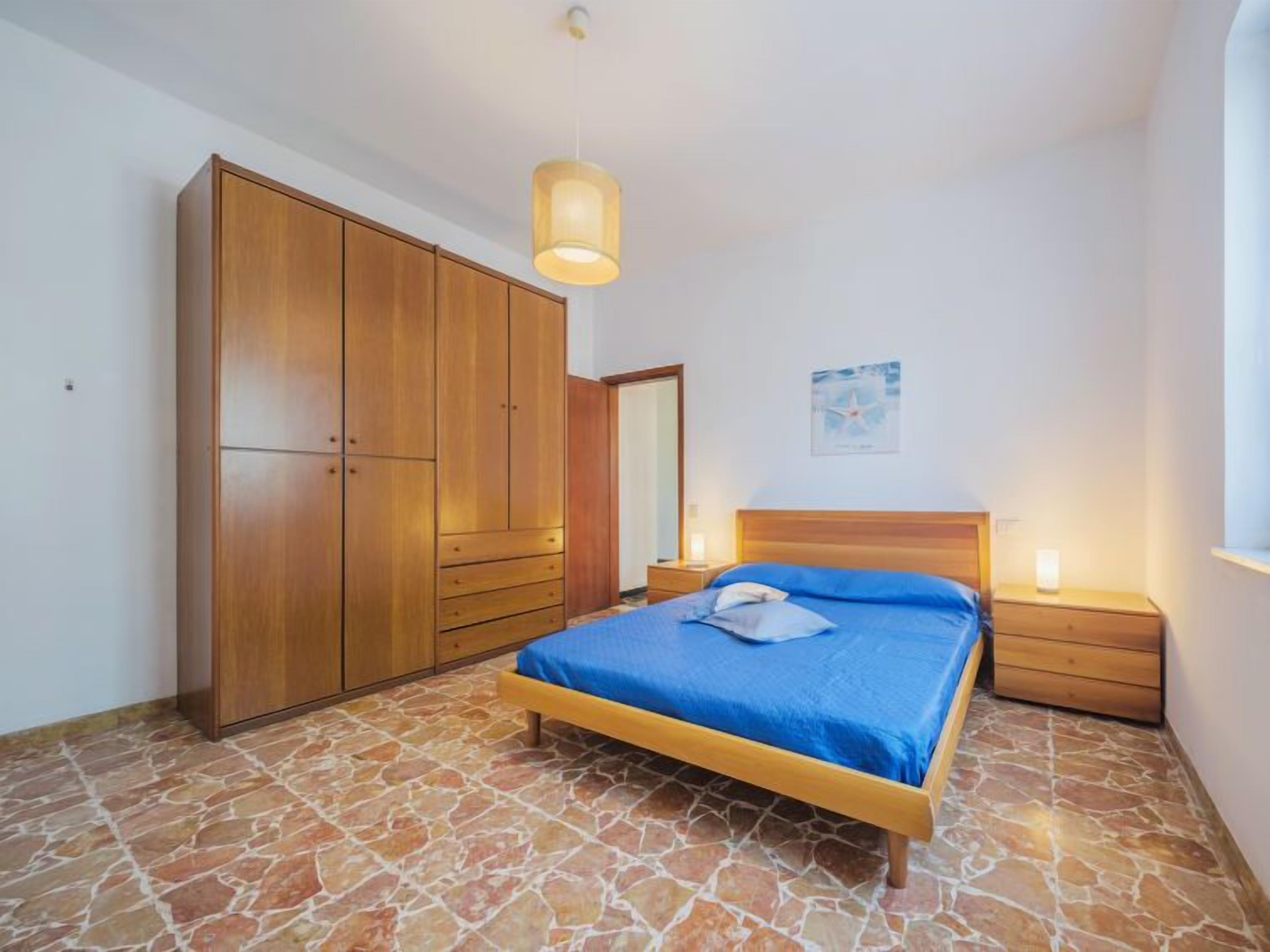 A bedroom with a bed, wooden wardrobe, stone flooring, and a nightstand with a lamp.