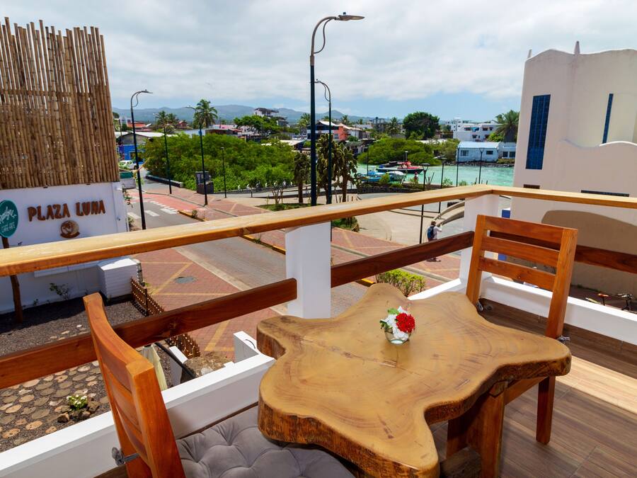 Una mesa y sillas de madera en un balcón exterior con vista a una playa y edificios al fondo.