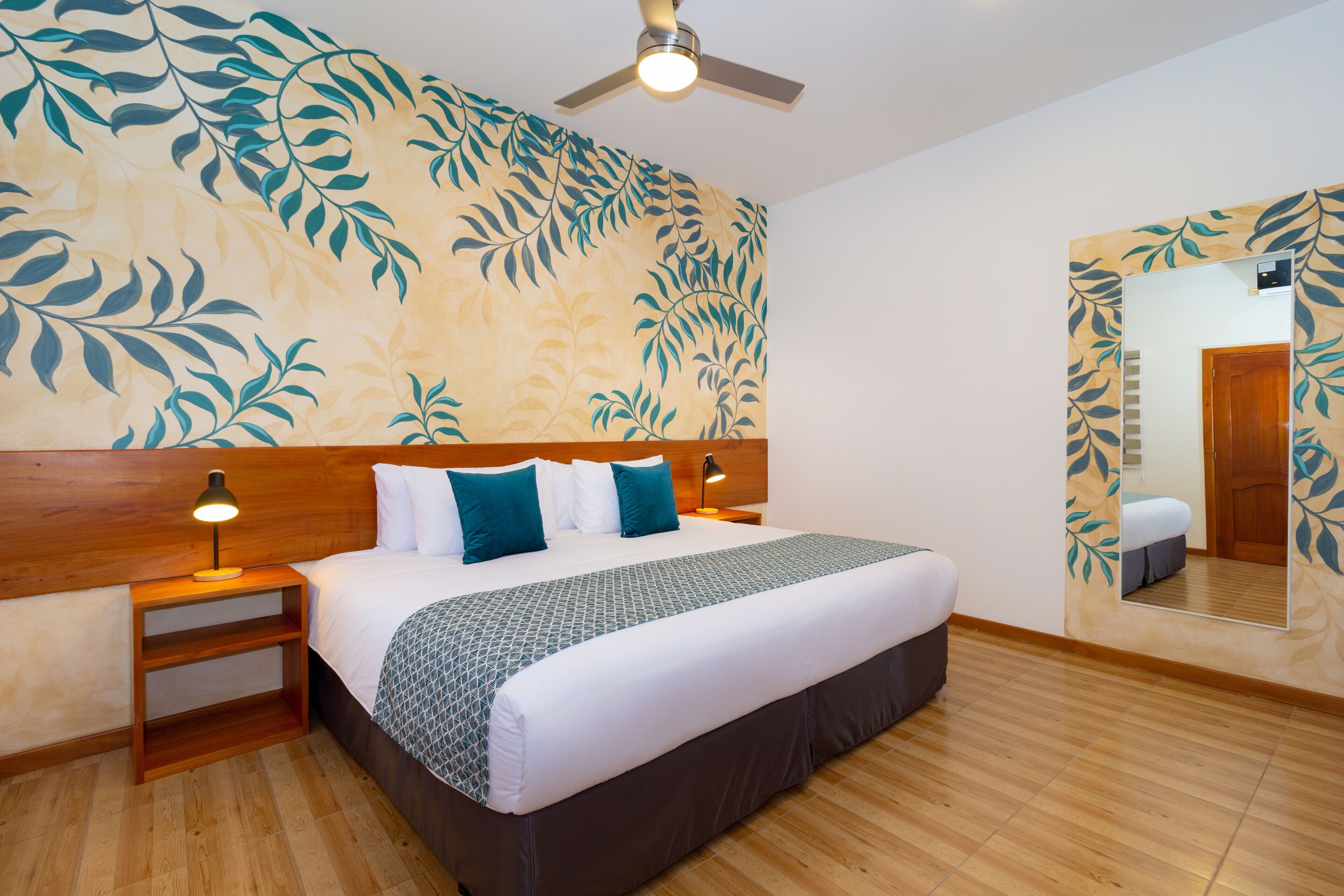 Une chambre d’hôtel avec un grand lit, une tête de lit en bois, un ventilateur de plafond, une table de chevet avec une lampe, un miroir et un plancher en bois.