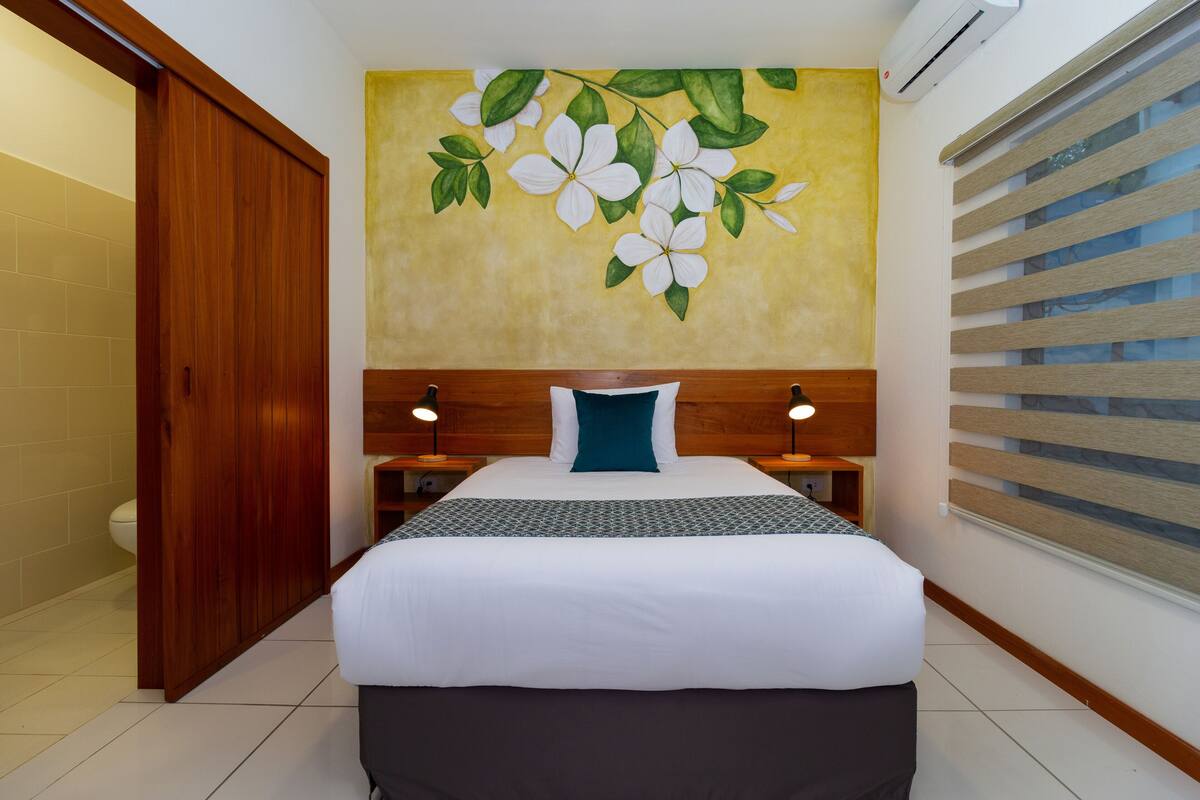 Une chambre d’hôtel avec un lit, une tête de lit en bois, un climatiseur fixé au mur et une fenêtre avec des stores.