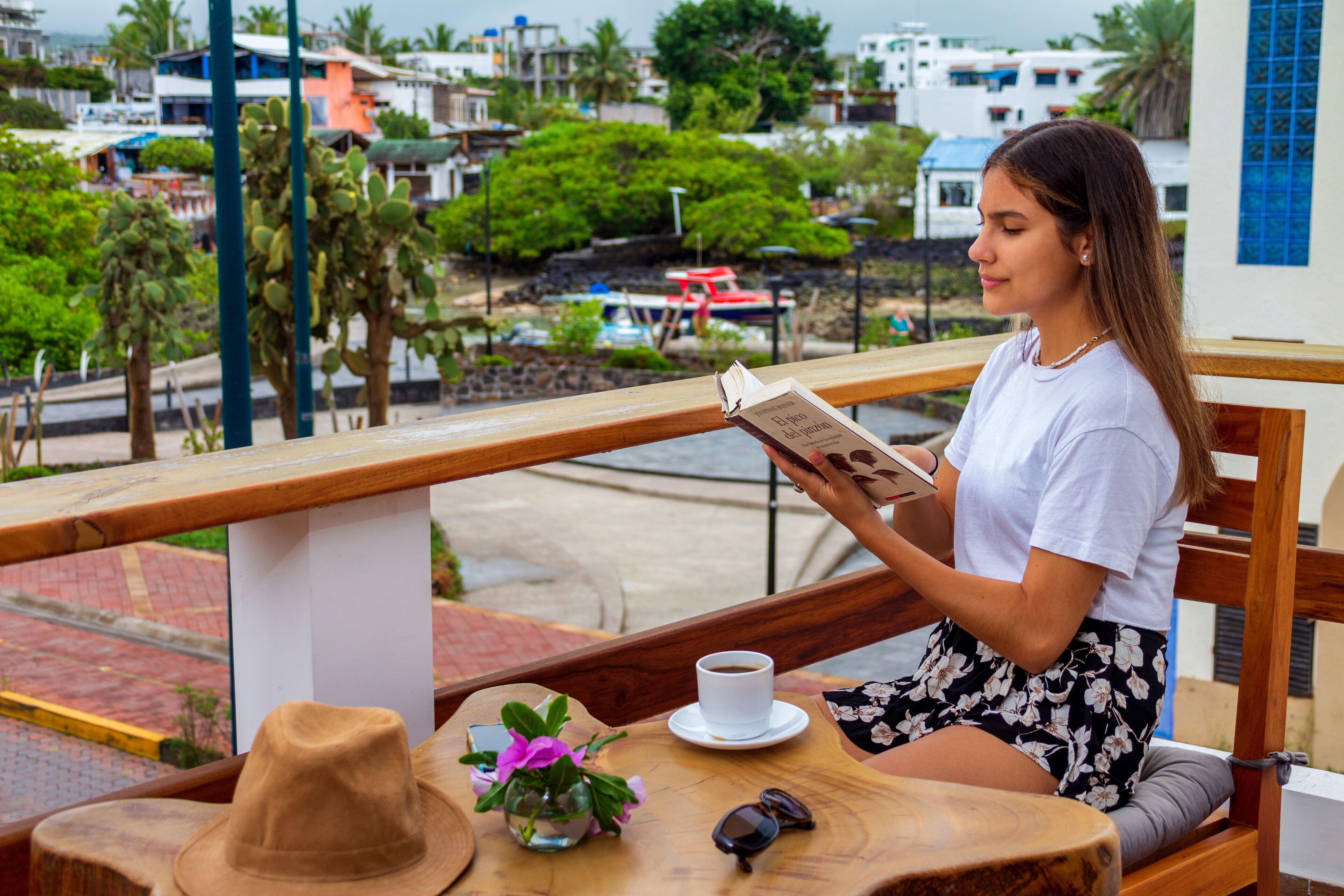 Une femme est assise à une table en bois, un livre ouvert devant elle, et elle lit. Sur la table, on aperçoit également une tasse de café, un chapeau et des lunettes de soleil.