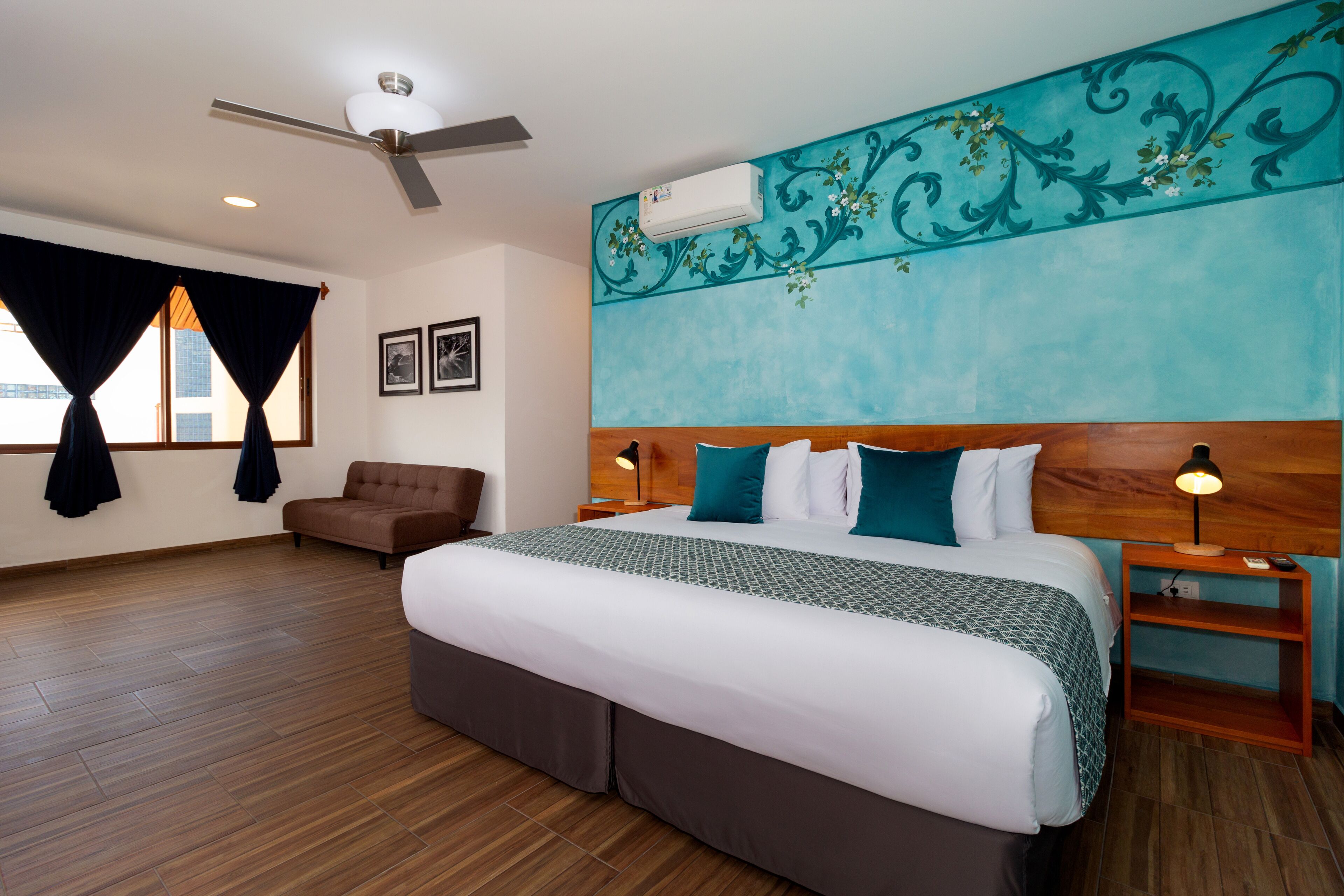 Een hotelkamer met een groot bed, een plafondventilator, een airconditioning, een houten hoofdeinde, een nachtkastje met een lamp en uitzicht op de stad door een raam met donkere gordijnen.