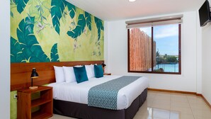 Habitación de hotel con una cama grande, una mesita de noche de madera, un mural en la pared, un ventanal con vistas a un cuerpo de agua y un televisor de pantalla plana montado en la pared.