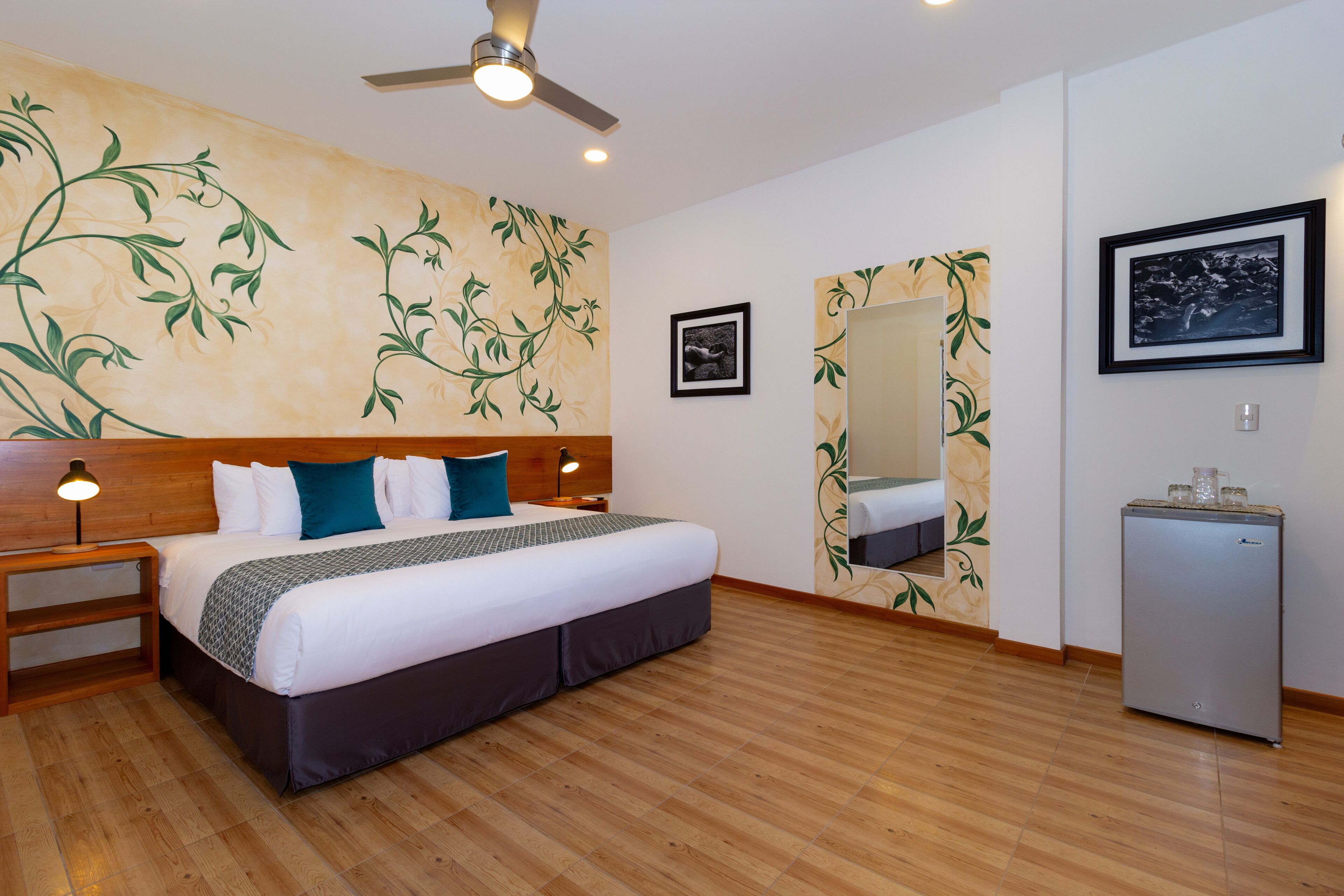 A hotel room with a wooden floor, a bed with white bedding and teal pillows, a nightstand with a lamp, a ceiling fan, a small refrigerator, and a wall with a floral wallpaper design.