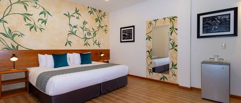 A hotel room with a wooden floor, a bed with white bedding and teal pillows, a nightstand with a lamp, a ceiling fan, a small refrigerator, and a wall with a floral wallpaper design.
