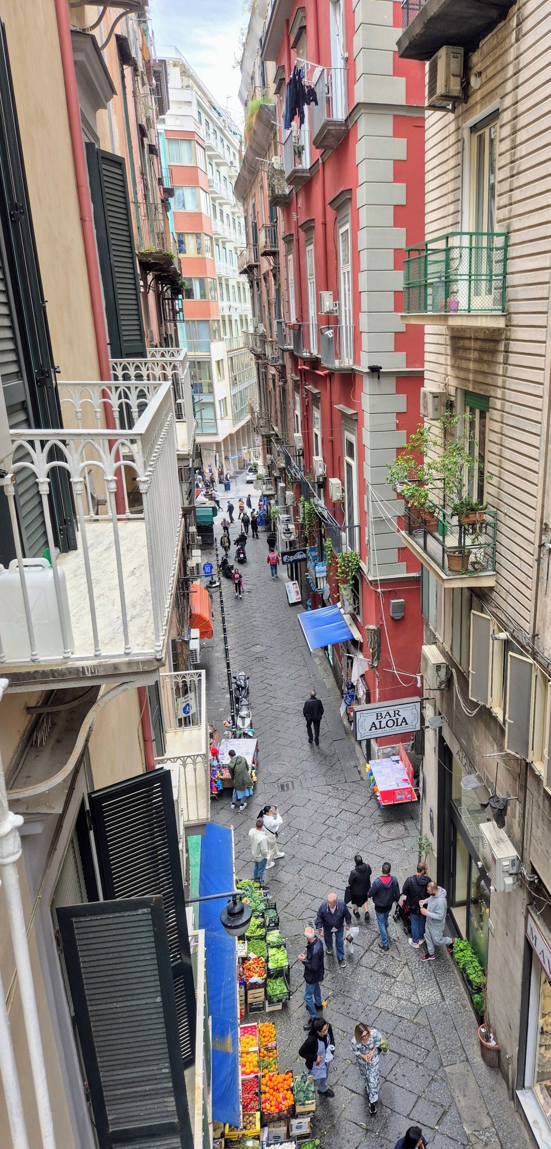 Una calle estrecha con balcones, un mercado de frutas y un bar.