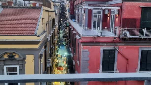 A narrow street with buildings on either side, a view of a hill with a castle-like structure in the distance, and a balcony with a railing.
