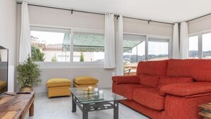 Un salon moderne avec un canapé rouge, une table basse en verre et une table d’appoint en bois. Une grande fenêtre est agrémentée de rideaux blancs, et on y trouve une plante en pot. La fenêtre offre une vue sur les bâtiments environnants.