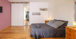 A bedroom with a wooden floor, a bed with grey bedding, a nightstand with a lamp, and a wardrobe with a mirror. - Domaine de la Bergerie en Drôme Provençale (SAUZET)