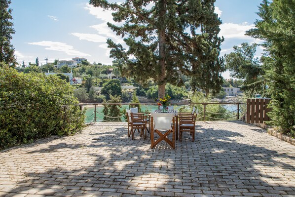 A paved outdoor patio with a table and chairs, surrounded by trees and overlooking a scenic view. - Grand Bleu Porto Heli Beachfront House (Ermionida)