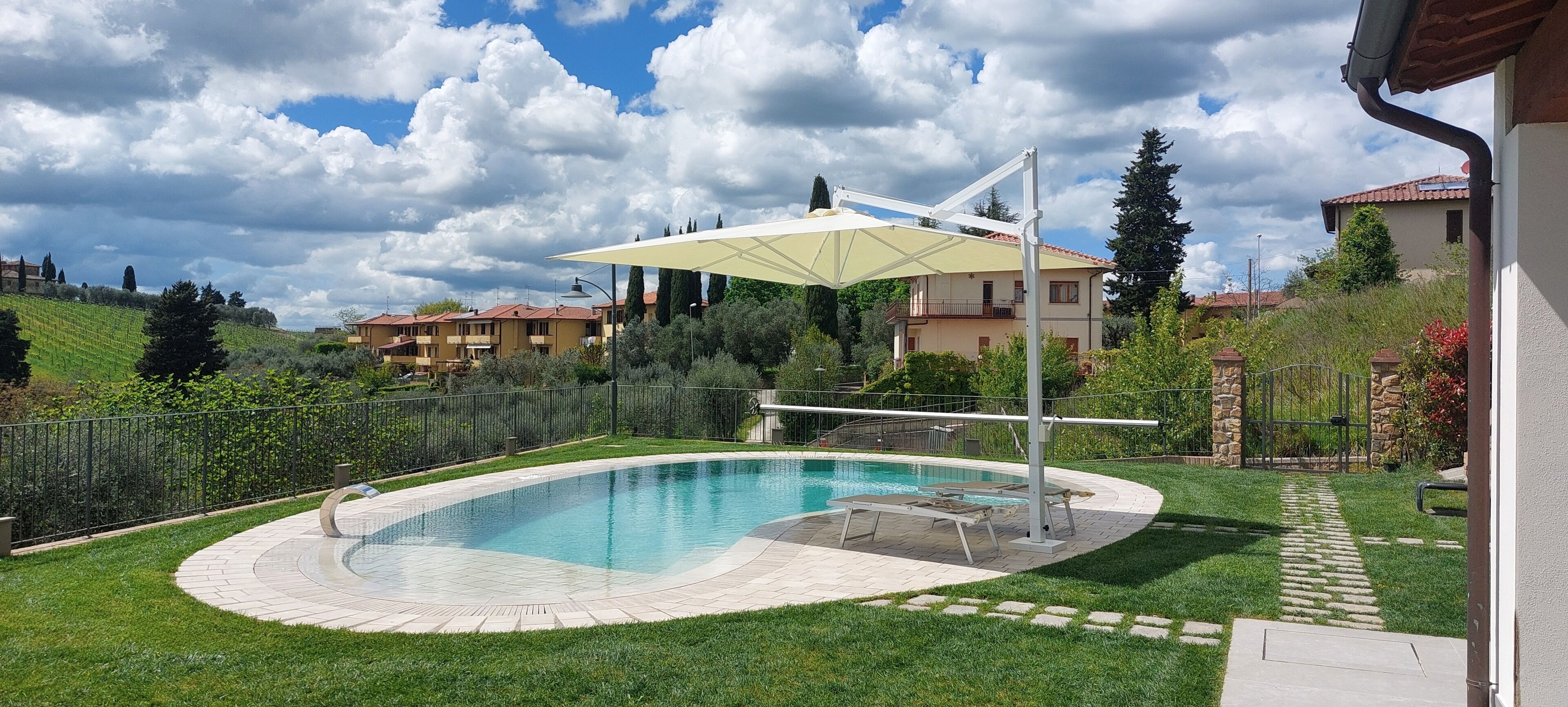 A swimming pool with a sun umbrella, surrounded by a paved deck and a stone border.