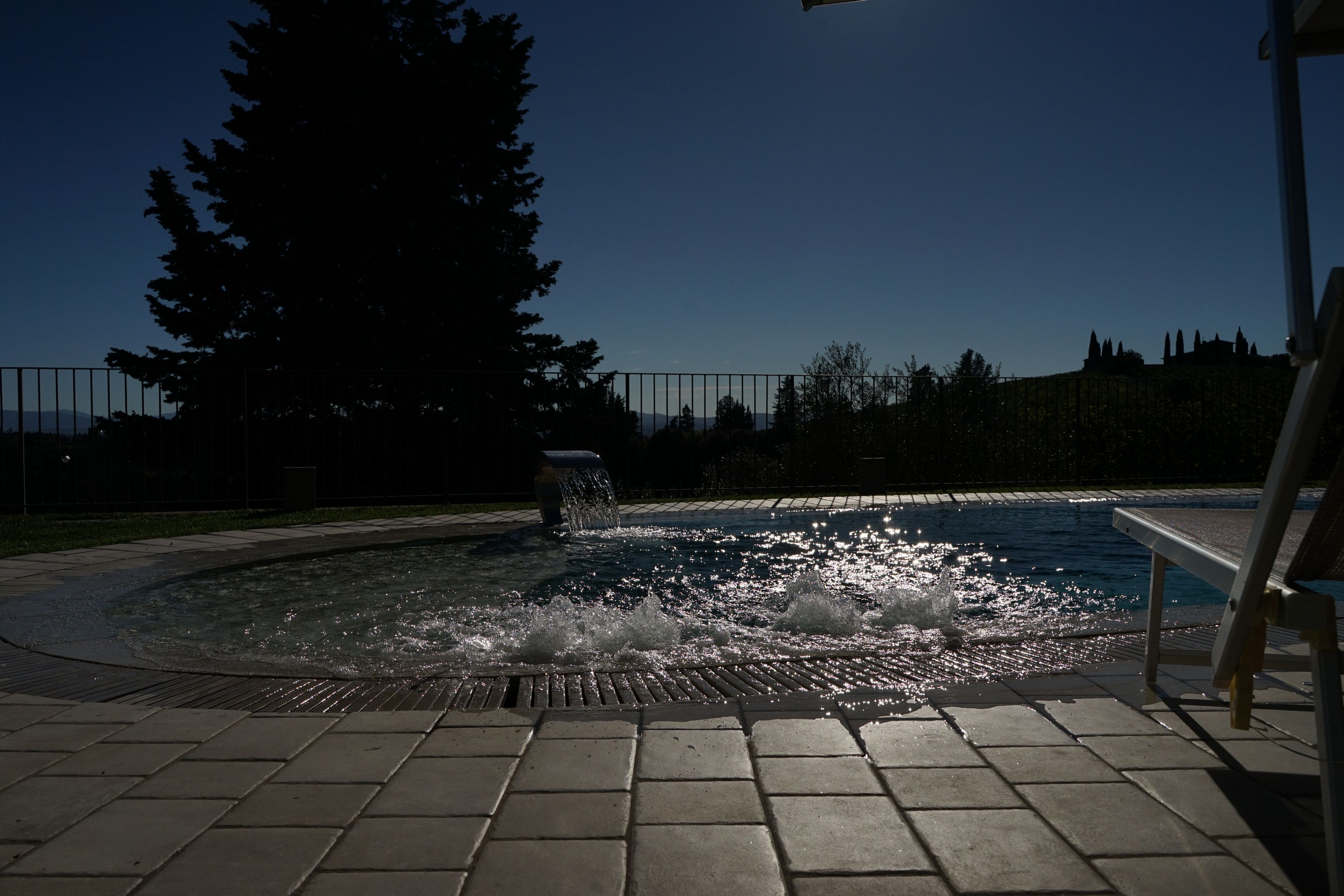 Ein Schwimmbecken mit einer Wasserspende in der Mitte, umgeben von einer gepflasterten Fläche und einem Zaun.