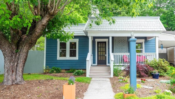 A two-story house with a front porch, a large tree, and a garden with various plants and flowers. - Artisan Gem - 2 BR / 1 Bath (Spruce Pine)