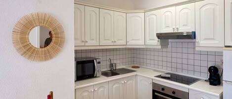A compact kitchen with white cabinets, a microwave, a stove, and a sink. There's a dining table with a fruit bowl and a mirror on the wall.