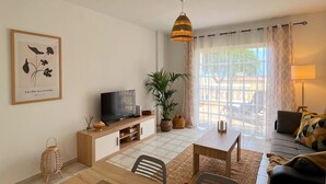 Ein Wohnzimmer mit Esstisch, Stühlen, Fernseher, Sofa und einem Fenster mit Vorhängen.