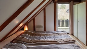 A bedroom with a sloped ceiling, wooden beams, a bed with striped bedding, a window with a view of a deck, and a wardrobe. - Rumpelstilzchen by Interhome (Frankenau)