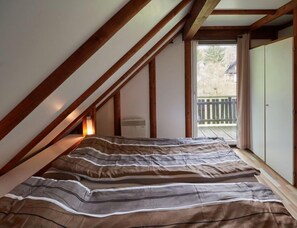 A bedroom with a sloped ceiling, wooden beams, a bed with striped bedding, a window with a view of a deck, and a wardrobe.