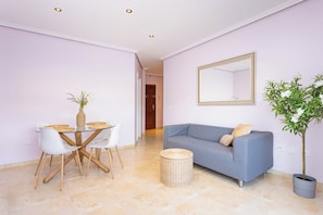 A modern living room with a grey sofa, a dining table with chairs, a round wicker side table, and a potted plant.