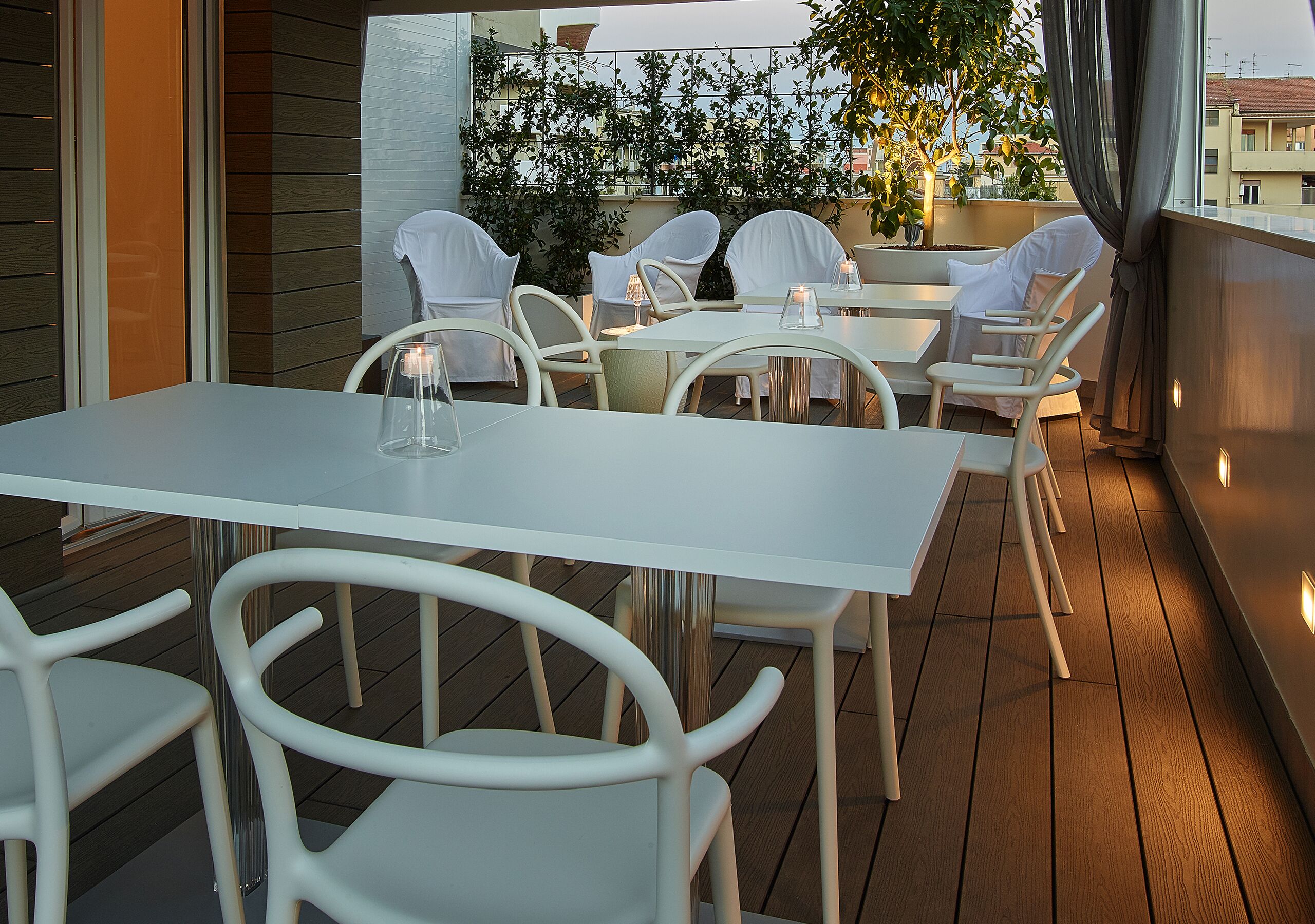Een terras met witte tafels en stoelen, een kaars op een tafel en gebouwen en bomen in de verte.