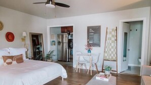 Une chambre avec un grand lit, une petite table et une chaise. Une porte donne accès à une cuisine et à une salle de bain.