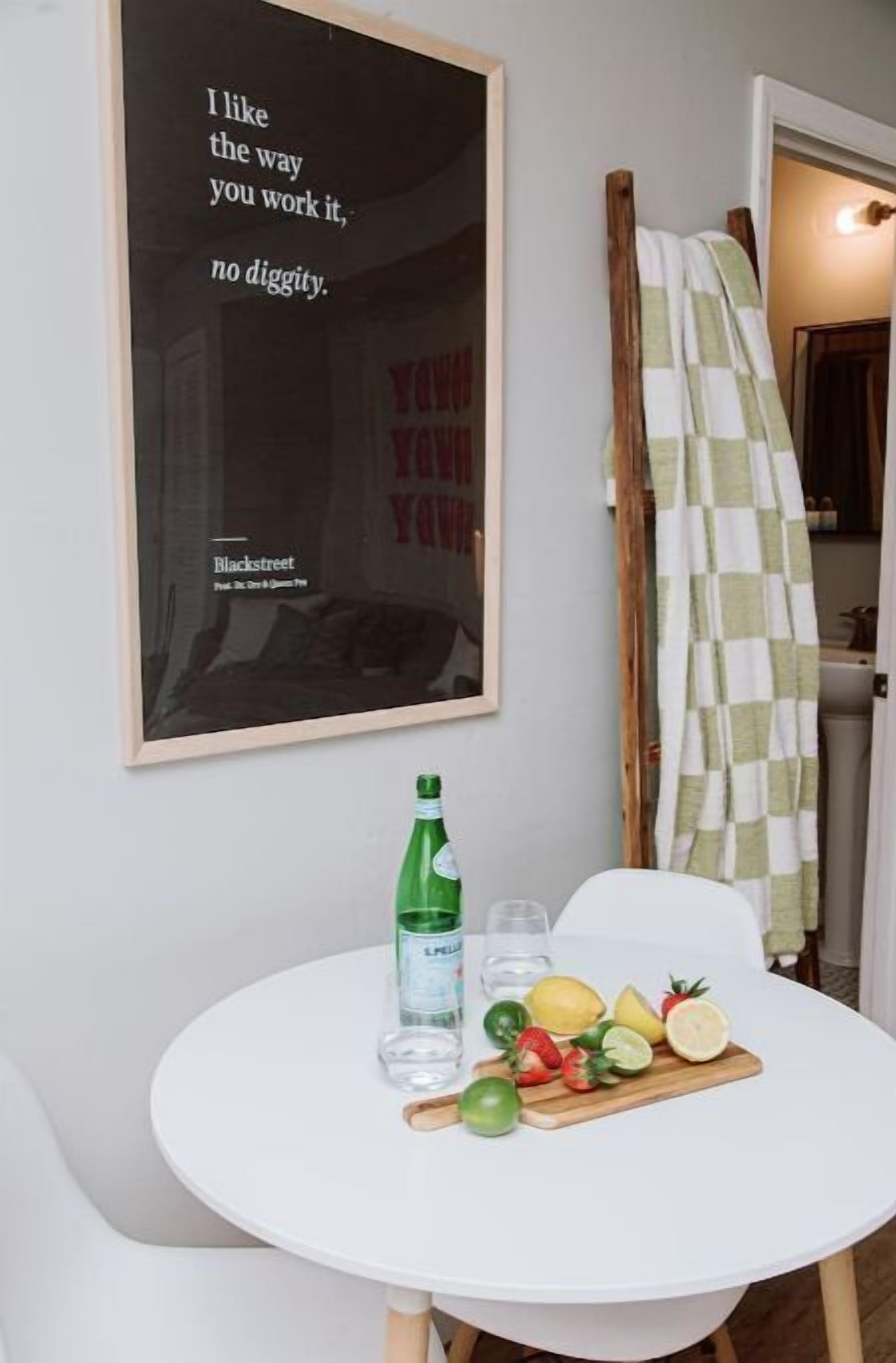 Un coin cuisine avec une table blanche, une bouteille d’eau, un verre et une planche à découper avec des fruits. Une affiche accrochée au mur porte l’inscription : « J’aime la façon dont tu t’y prends, c’est parfait. »