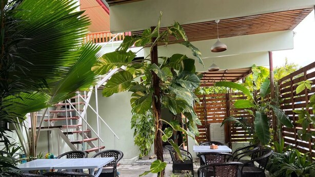 An outdoor patio with tables and chairs, surrounded by lush greenery and a wooden pergola.