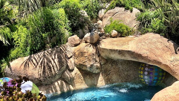 Ein Schwimmbecken mit einem Wasserfall-Element aus Felsen, umgeben von üppiger Vegetation.
