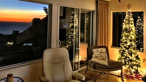 Ein Wohnzimmer mit großem Fenster, einem Weihnachtsbaum und Blick auf das Meer bei Sonnenuntergang.